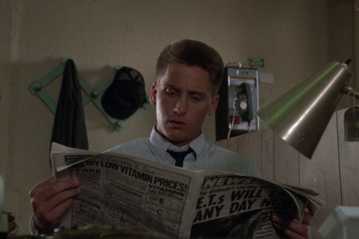 Emilio Estevez reads a newspaper with text about aliens coming and low vitamin prices in Repo Man