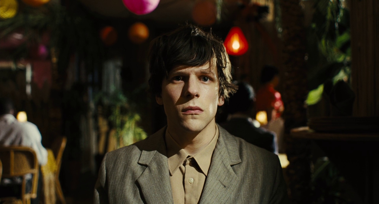 Jesse Eisenberg looks solemn and stares at the camera while standing in what looks like a restaurant in The Double