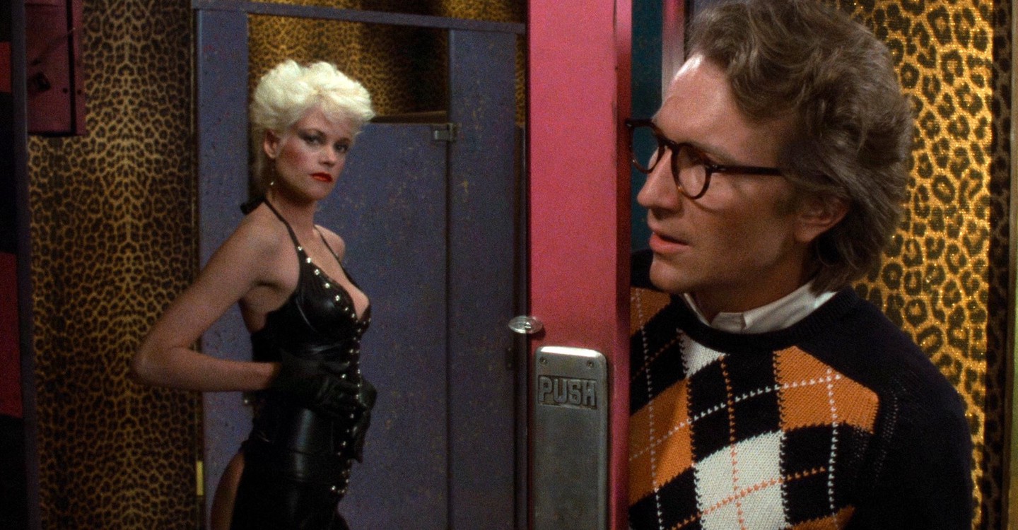 A man in a black, white, and orange argyle sweater and wearing glasses stares at a blond-haired woman in leather outfit reflected in a mirror in Body Double.