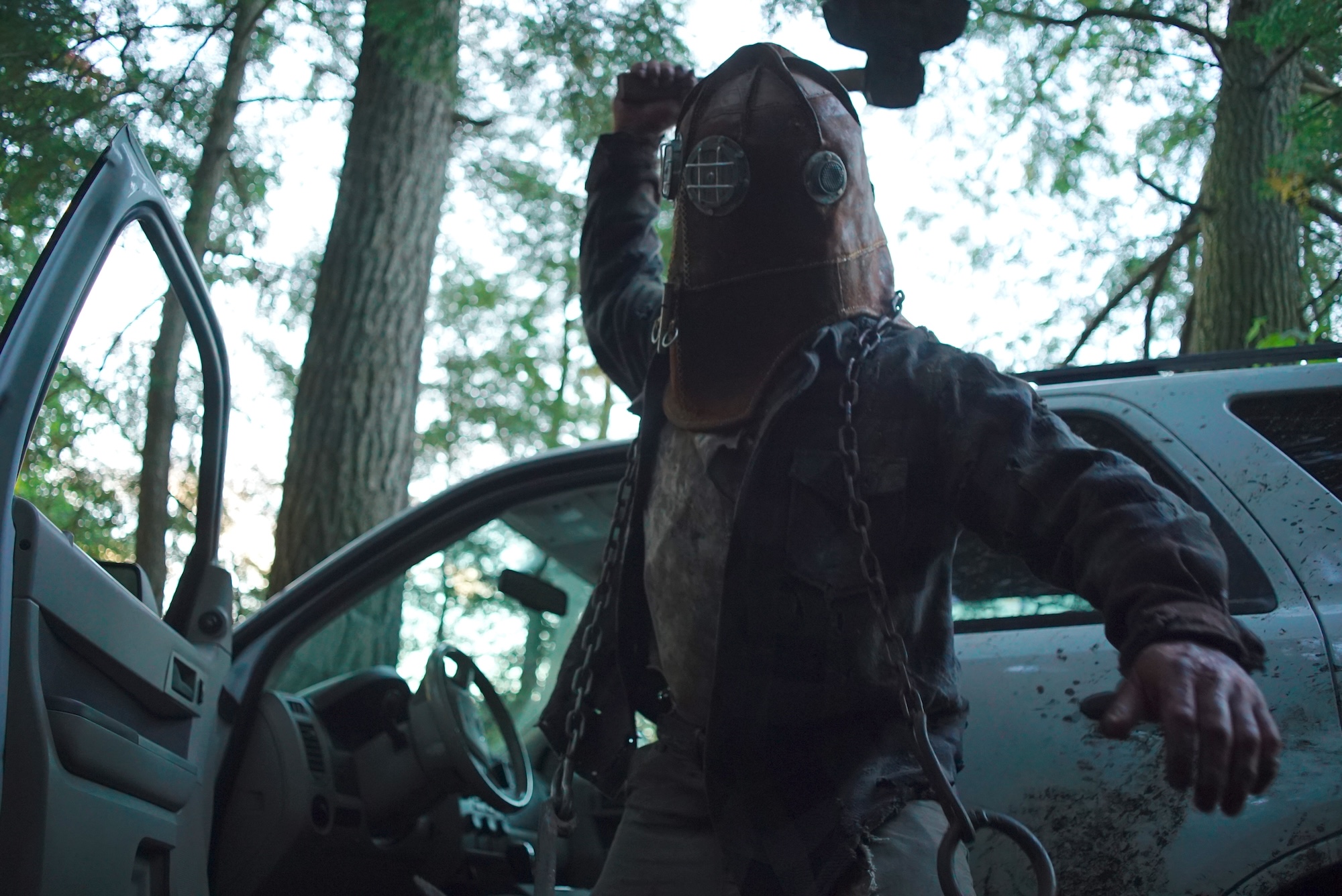 A hooded figure standing in front of a dirty white four-door van raises an axe to swing. They’ve got chains draped around their shoulders and appear to be in the middle of the woods. (In a Violent Nature)