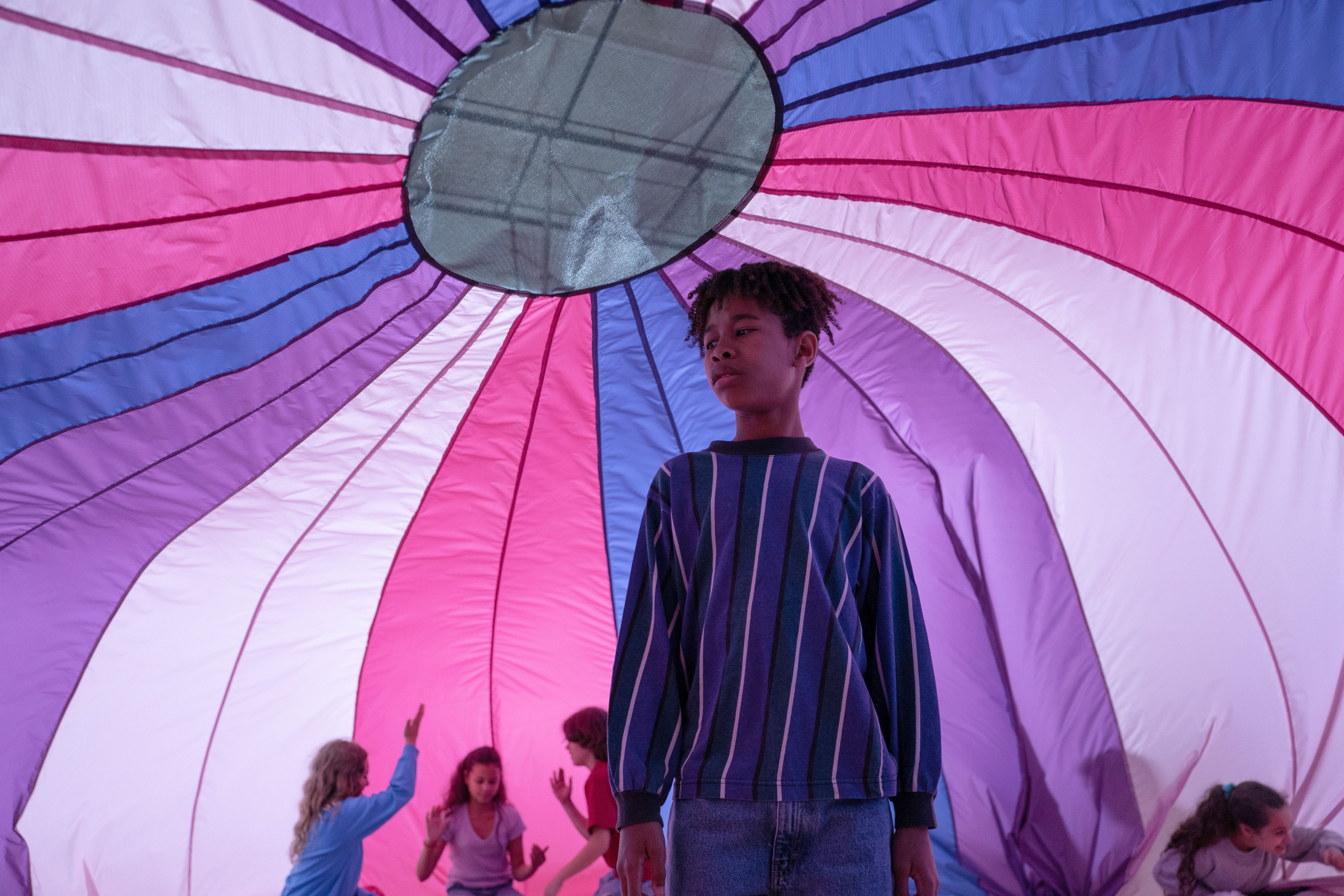 Young Owen (Ian Foreman) stands with classmates under a huge gym-class parachute in the bisexual-flag colors of pink, blue, and purple in Jane Schoenbrun’s movie I Saw the TV Glow