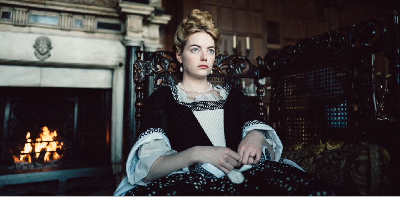 Emma Stone in THE FAVOURITE, in fancy dress in a fancy chair and sitting in front of a fancy fireplace