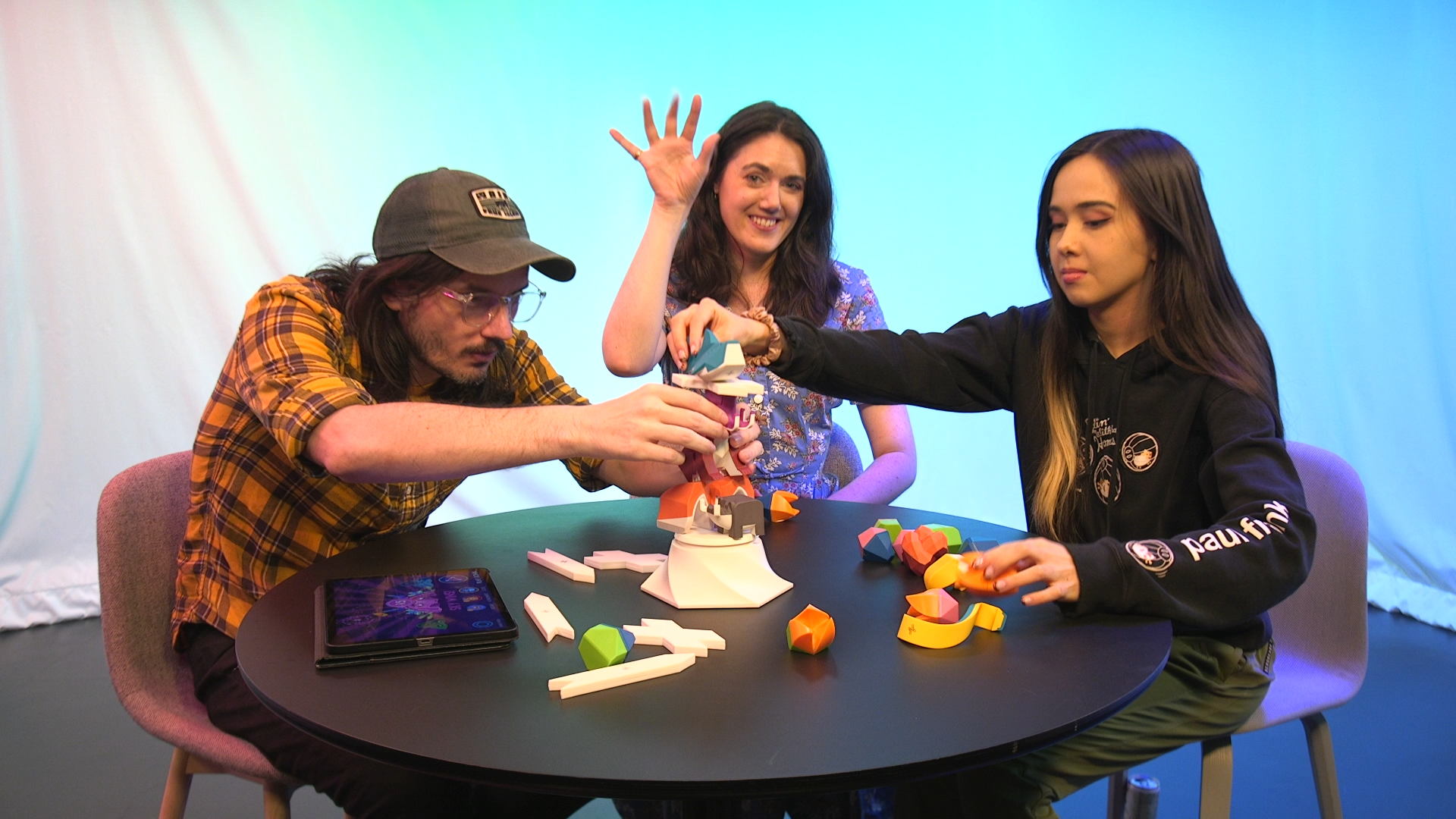 Three people sitting at a table with a colorful backdrop. Two players are stacking animal shaped blocks in the game Beasts of Balance while one waves at the camera.